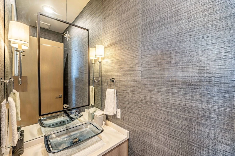 The elegant powder room is perfect for guests. Featuring high end appliances, custom glass sink with beveled glass mirror and dark grey linen wallpape